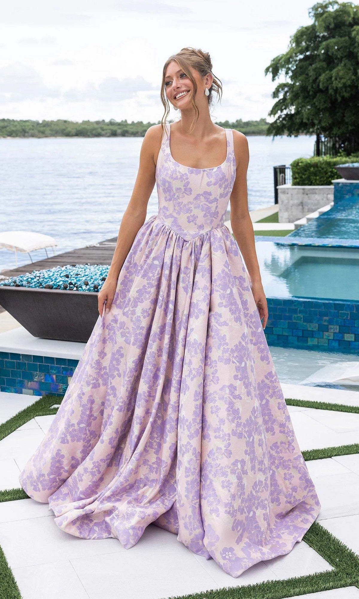 A woman stands outdoors by a pool, smiling and holding the sides of her dress while wearing the Portia and Scarlett XO185 long formal dress in lilac/pink.